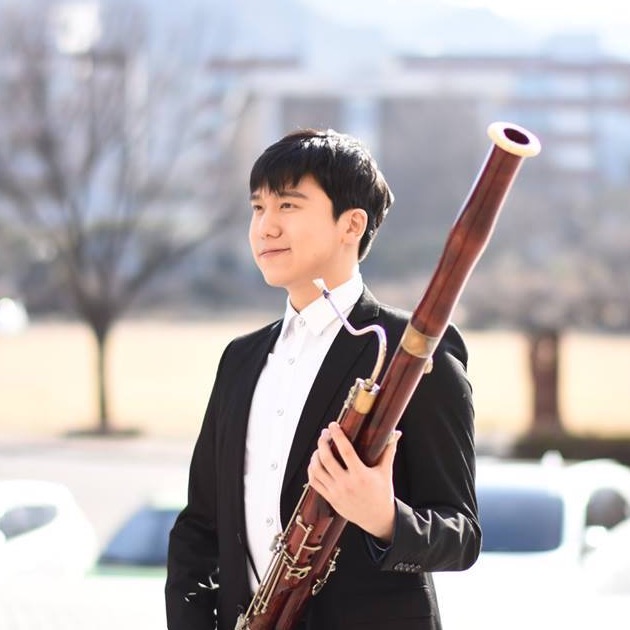 Profile Picture of Minkyu Kim: A man with short dark hair, smiling gently, stands outdoors in a black suit with a white shirt, holding a bassoon vertically in front of him. He appears to be a musician posed for a portrait with a clear sky and indistinct buildings in the background.